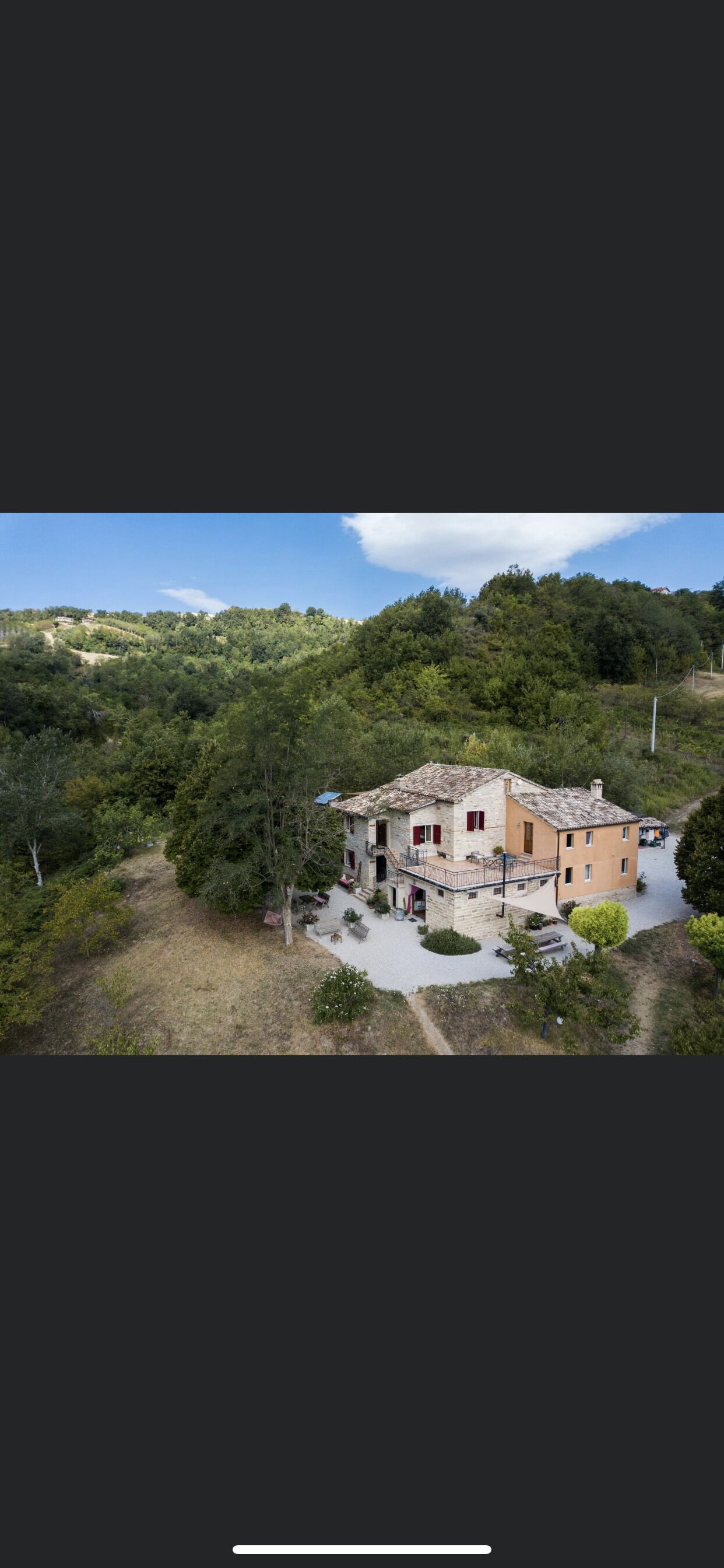 Una casa in pietra a due piani con un balcone, immersa in un giardino e alberi, situata su un pendio.