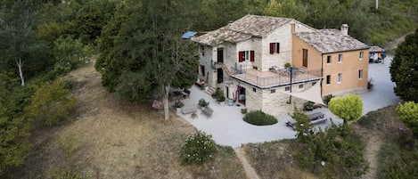 A two-story stone house with a balcony, surrounded by a garden and trees, located on a hillside.