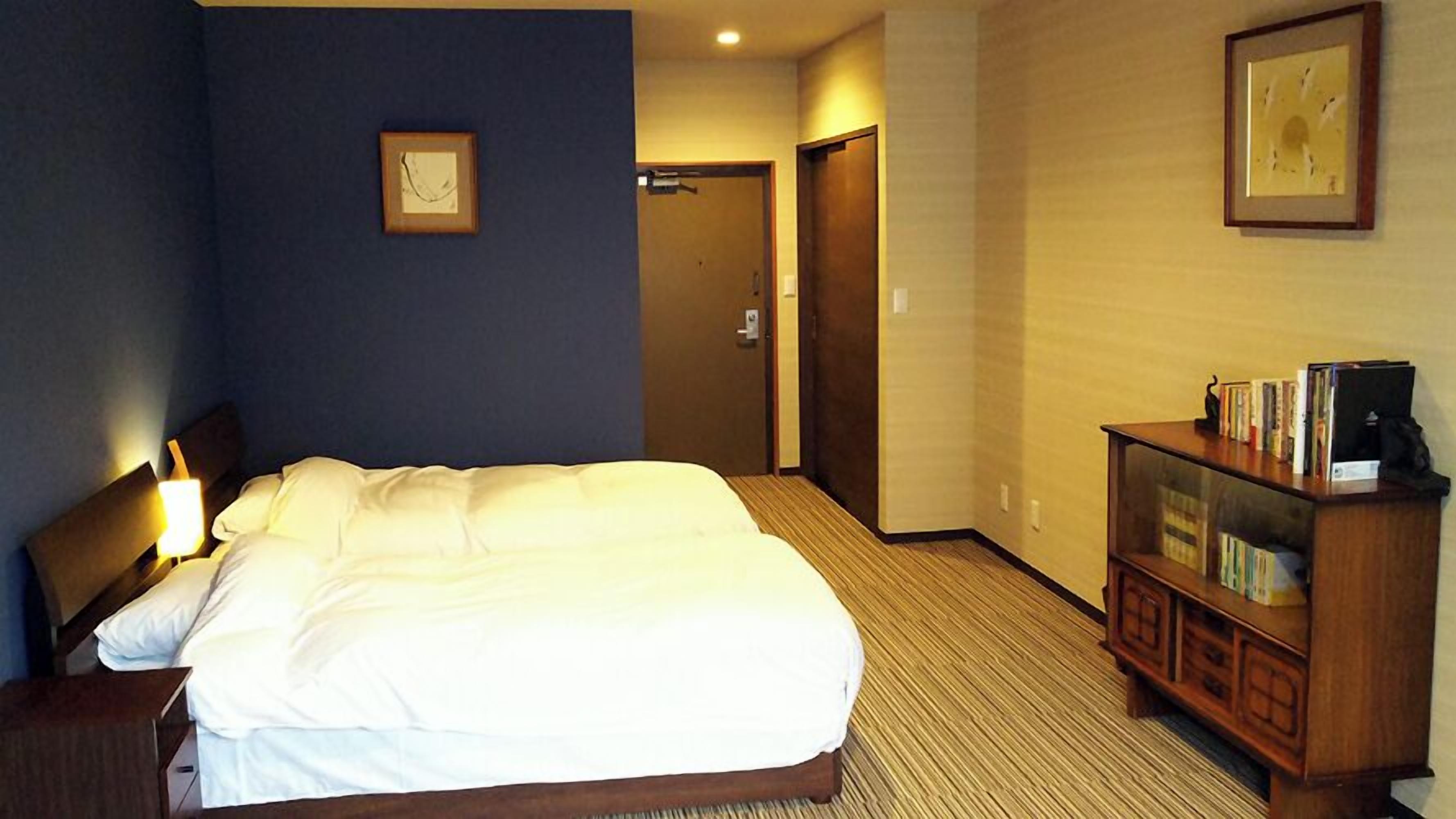 Une chambre d’hôtel avec un lit, une table de chevet, une lampe, une armoire en bois remplie de livres et une porte donnant sur une autre pièce.