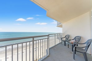 A balcony with a view of the beach and ocean, two chairs, and a table.