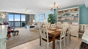 A spacious living area with a dining table, white chairs, a chandelier, and a bookshelf. - Ocean Bay Club 1508-Oceanfront 3BR-Great Amenities (North Myrtle Beach)