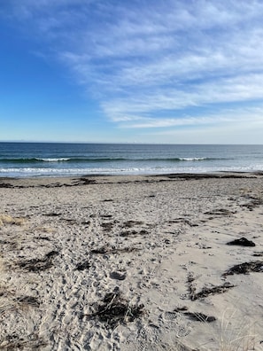 En strand med tydelige fodspor og tang, der fører ud til havet under en klar himmel.