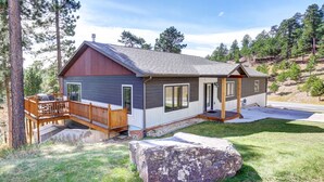 Una cabaña moderna con terraza, una roca grande al frente y un bosque de fondo.