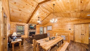 A cozy cabin interior with a wooden dining table, a fireplace, a sofa, and a television.