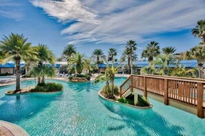 A resort pool area with multiple water features, palm trees, and a wooden bridge. -  Destin's Salty Beach Paradise! (Destin)