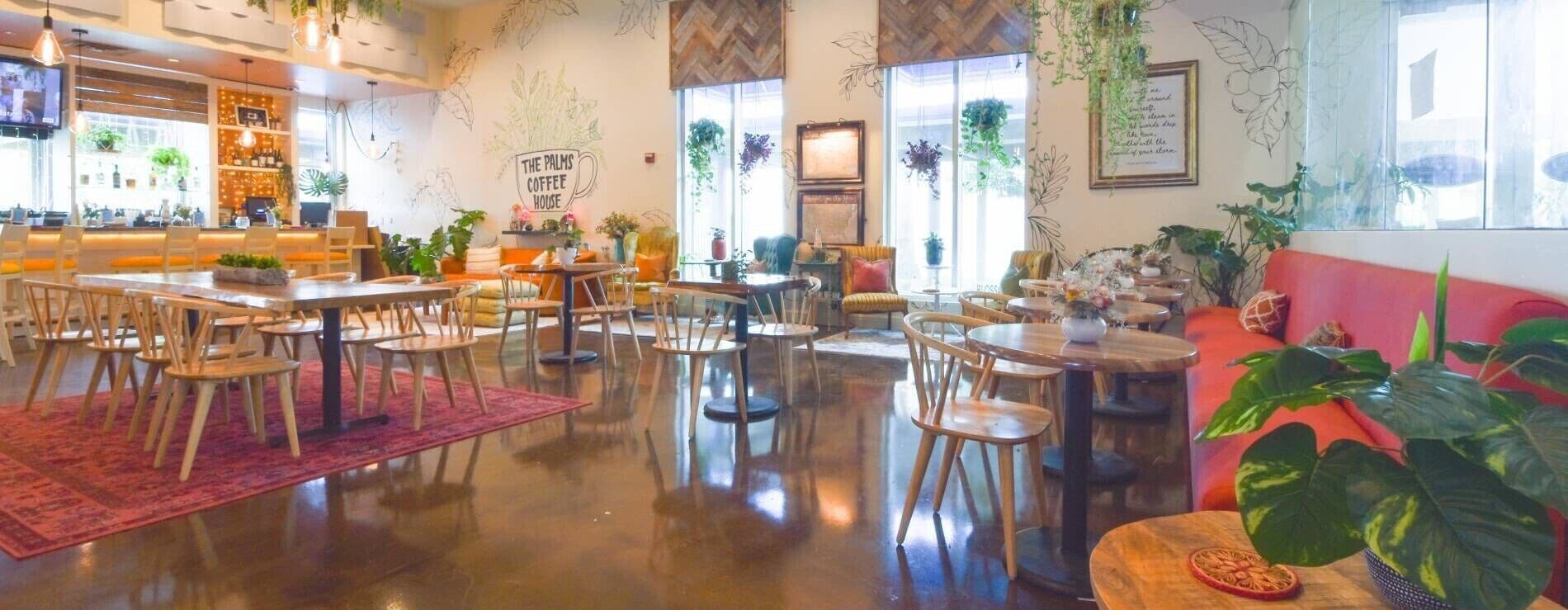 A spacious coffee shop with multiple seating areas, including tables and chairs, and a variety of plants.