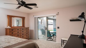 A bedroom with a bed, a dresser, a desk with a lamp, a ceiling fan, and a sliding glass door leading to a patio with chairs and a table. - Cliffrose pool home with RV/Boat parking (Lake Havasu City)
