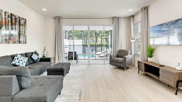 A modern living room with grey sofas, a coffee table, a TV stand, and a large window overlooking a pool area. - Edna's floridian hidden retreat (Clermont)
