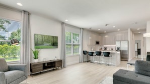 A modern living room with a grey sofa, a dining area with a bar, and a kitchen with a stainless steel refrigerator. - Edna's floridian hidden retreat (Clermont)