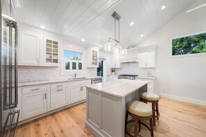 A modern kitchen with white cabinetry, a central island with stools, and a large window providing natural light. - Sandals D | Luxury Beachfront Living on Sanibel Island (Sanibel)