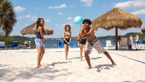 Une scène de plage où l’on voit des gens jouer au volleyball, des parasols en chaume et des chaises longues.