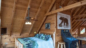 Cozy cabin-style room with a high ceiling and exposed beams, featuring a plush bed and a charming table and chair.