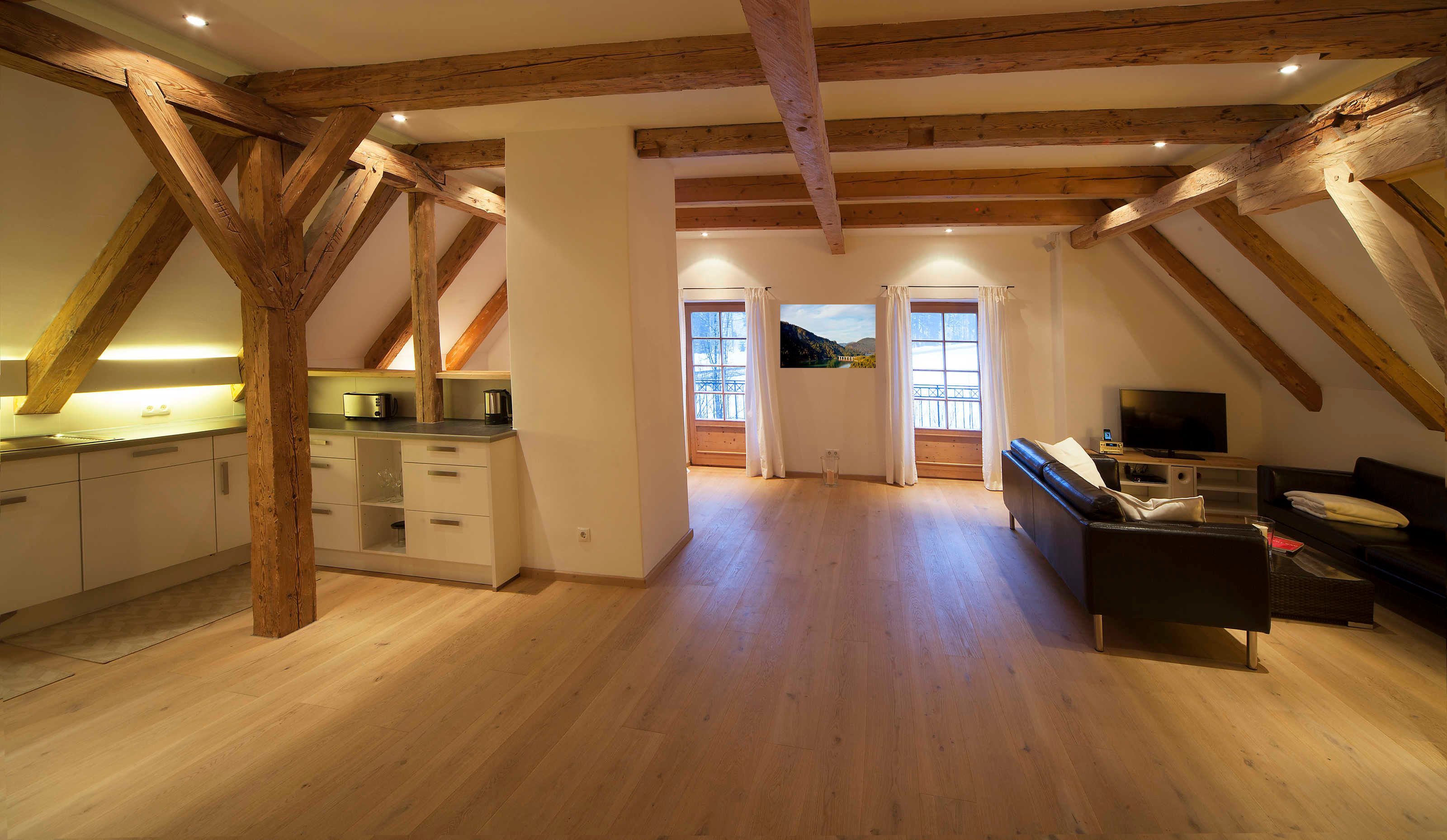 A spacious loft with exposed wooden beams, a kitchen with modern appliances, a living area with a sofa and TV, and a large window with a scenic view.