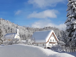 A snow-covered house with a white roof and a church with a cross on top, surrounded by snow-covered trees.