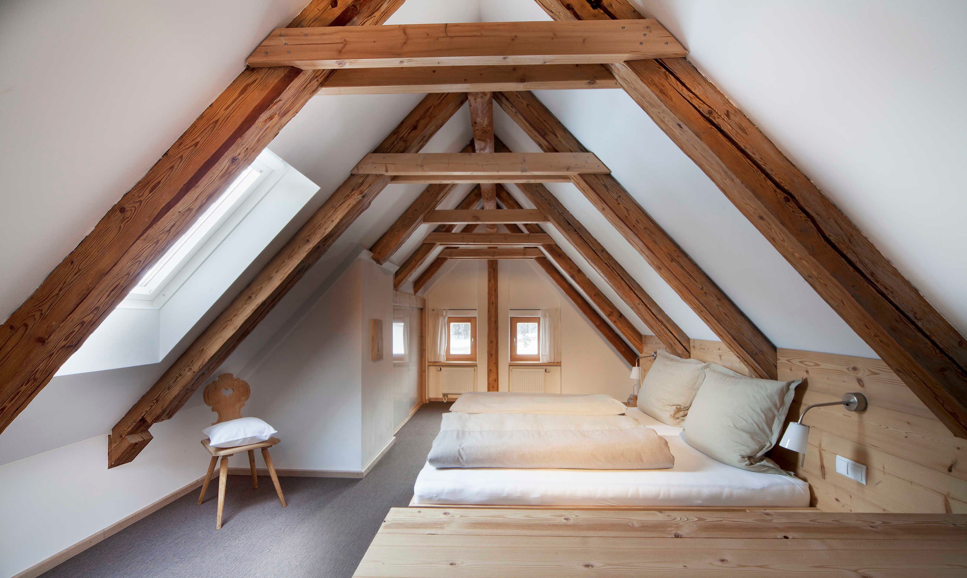A loft bedroom with a sloped ceiling, wooden beams, a bed, a chair, and a small table.