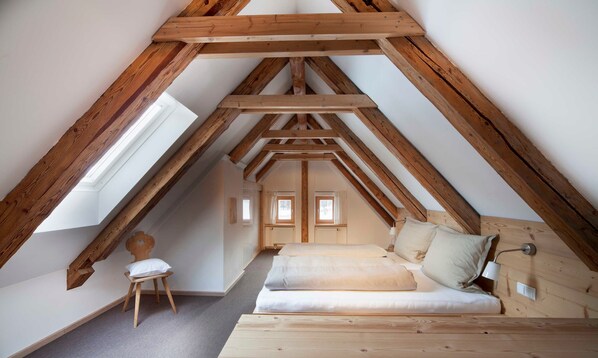 A loft bedroom with a sloped ceiling, wooden beams, a bed, a chair, and a small table.