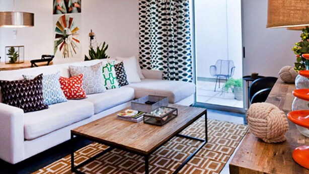 Un salon moderne avec un canapé blanc, des coussins à motifs, une table basse en bois et une vue sur l’extérieur grâce à une porte vitrée.