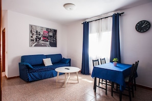Une pièce avec un canapé bleu, une table ronde, une table à manger avec des chaises, une horloge au mur et une fenêtre avec des rideaux.