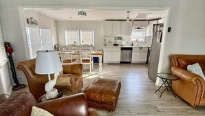 A spacious living room with a brown leather sofa, a dining table with chairs, and a kitchen area in the background.