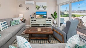 A living room with a grey sofa, a wooden coffee table, and a grey armchair. There is a white cabinet with glass doors, a plant, and a painting of a beach scene. - Bayfront with great patio! (San Diego)