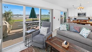A spacious living area with a sliding glass door leading to a patio, a grey sofa with patterned cushions, a wooden coffee table, and a view of boats and greenery outside. - Bayfront with great patio! (San Diego)