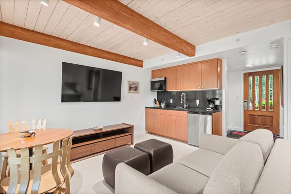 A modern living room with a sofa, a dining table with chairs, a kitchen area with appliances, and a TV mounted on the wall.