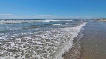 Una platja amb ones que brollen suaument a la riba, cel blau i edificis al lluny.