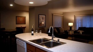 A modern kitchen with a double sink, a dishwasher, and a dining area with a table and chairs. There is a sofa and a lamp in the living area. - 5th Steele Place, LLC - Downtown Algoma Condos: Overnight or Extended Stays (Algoma)