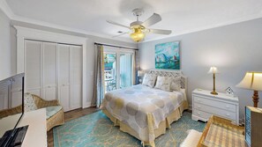Ein Schlafzimmer mit Bett, Deckenventilator, Nachttisch, Lampe, einem Gemälde und Blick ins Freie.