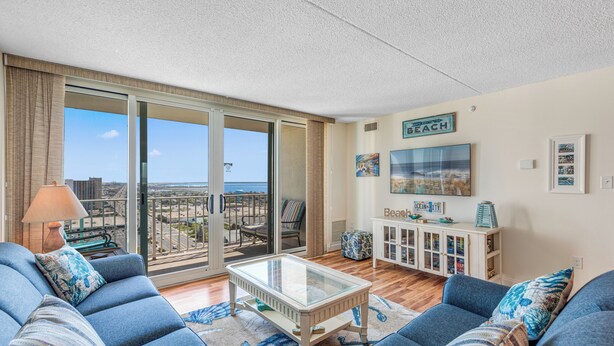 A living room with a glass-top coffee table, blue sofas, and a view of the beach through large sliding glass doors. - The Quay 2102 by Seaside Vacations & Sales (Ocean City)