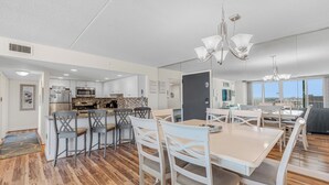 A modern kitchen with white cabinets, stainless steel appliances, and a dining area with white chairs and a large table. - The Quay 2102 by Seaside Vacations & Sales (Ocean City)