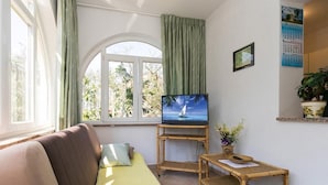 Een woonkamer met een bank, een televisie op een meubel, een klein tafeltje en een raam met gordijnen.