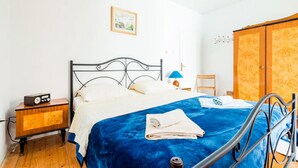 A bedroom with a bed, a wooden nightstand, a radio, a blue blanket, a white pillow, a wooden wardrobe, and a small table with a lamp. - Guesthouse Home Sweet Home - Double Room with Shared Terrace (Dubrovnik)