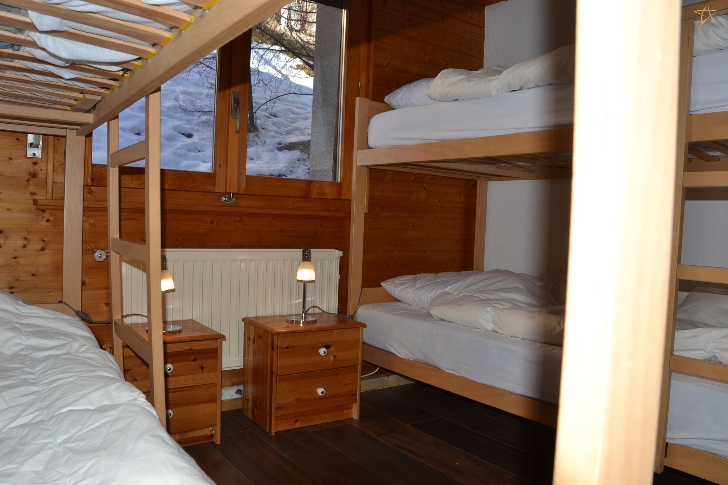A wooden cabin room with bunk beds, a small wooden nightstand with drawers, a window with a view of a snowy landscape, and a radiator under the window.
