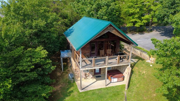 A wooden cabin with a green roof, a covered porch, and a stone foundation, surrounded by dense green trees. - A Painted Pony - Cabin (Sevierville)