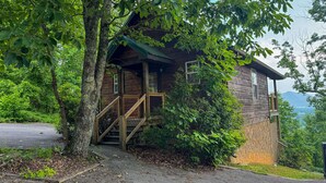 A rustic cabin with a wooden staircase leading to a porch, surrounded by lush greenery and a mountain backdrop. - Angel's View By Volunteer Cabin Rentals - Cabin In The Smokies (Sevierville)
