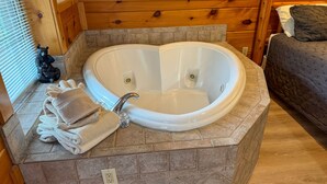A heart-shaped hot tub in a wooden cabin bathroom with a stone countertop, towels, and a bear statue. - Angel's View By Volunteer Cabin Rentals - Cabin In The Smokies (Sevierville)