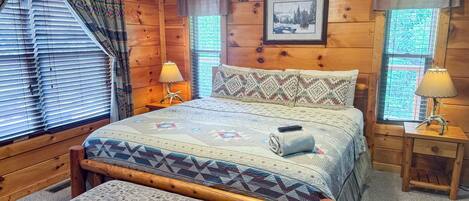 A wooden cabin bedroom with a bed, two bedside tables, a bench, and a window with curtains.