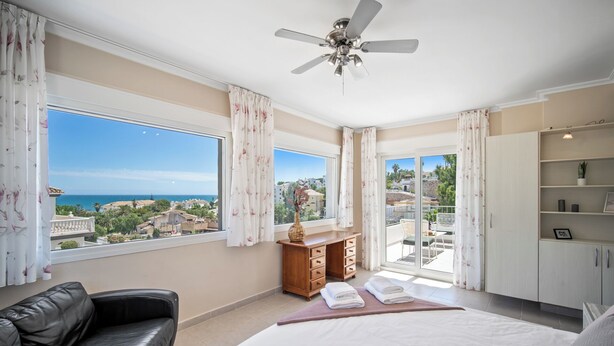 A bedroom with a bed, a wooden dresser, a black leather sofa, and a ceiling fan. There are large windows offering a view of the sea and buildings. - Villa Jimena by Mabiente Properties by Mabiente Properties (Mijas)