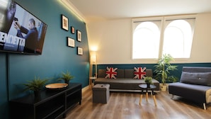 Ein Wohnzimmer mit einer Couch, einem an der Wand montierten Fernseher, einem kleinen Tisch mit einer Pflanze und einem Fenster mit rundbogigem oberen Rand.