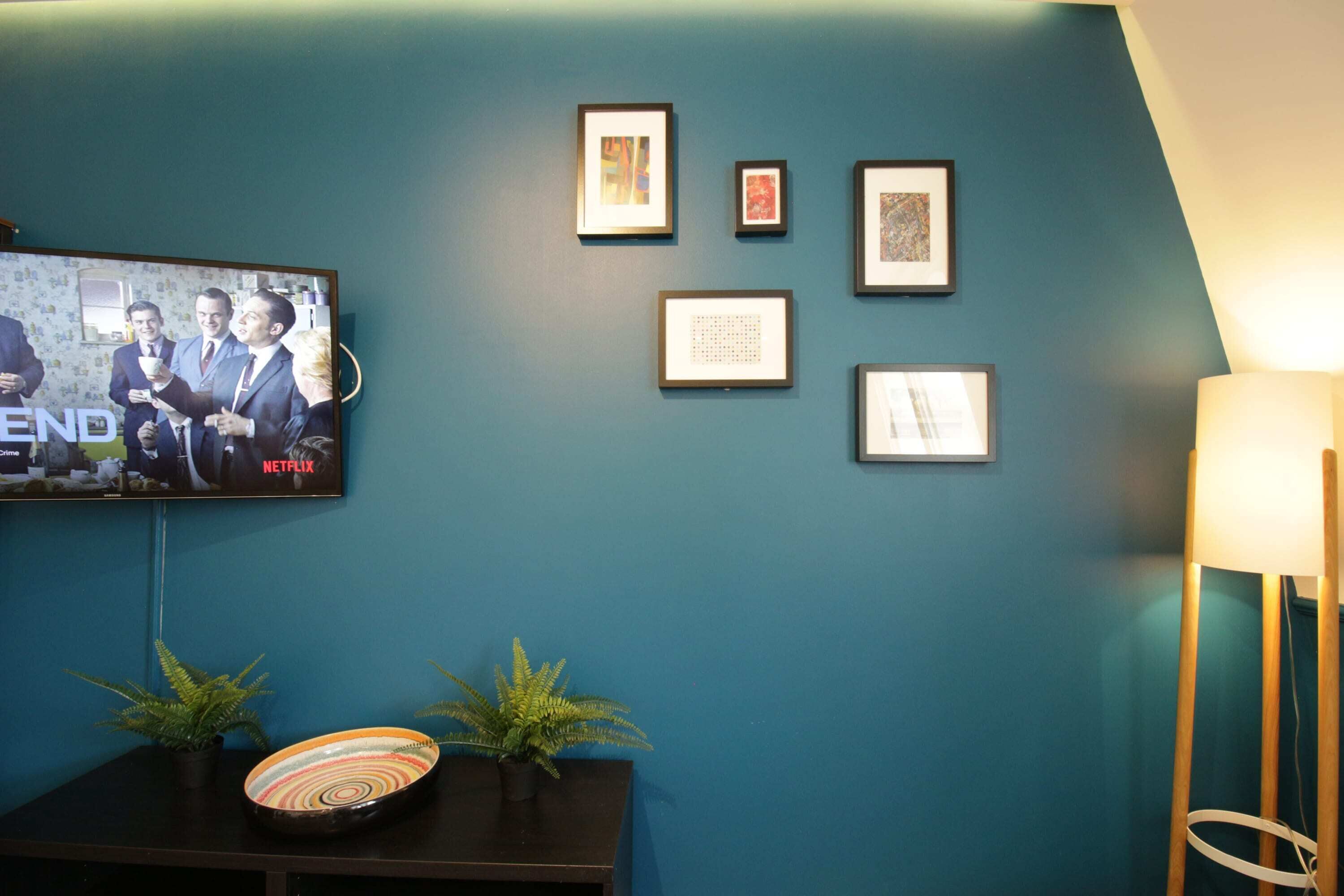 A room with a teal wall, a flat-screen TV displaying a show, framed pictures, a small table with decorative items, and a floor lamp.