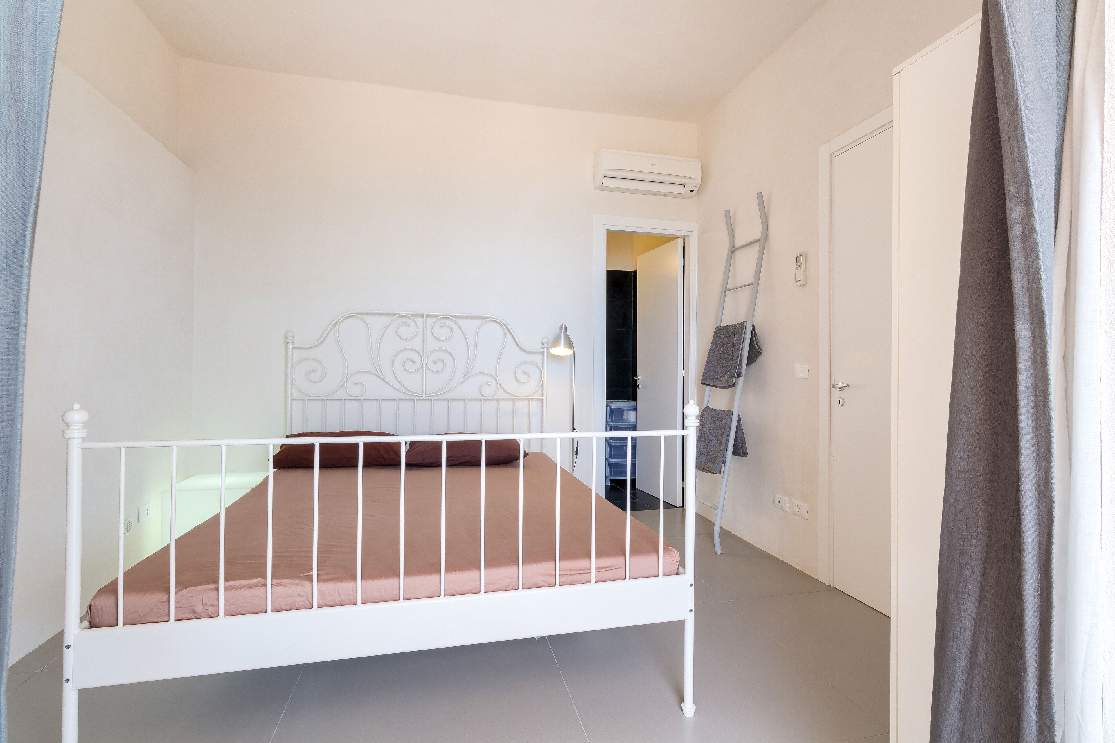 Une chambre avec un lit simple, dotée d’une tête de lit en métal blanche, d’un matelas marron et d’un mur blanc. On y trouve une porte qui mène à une autre pièce, ainsi qu’une échelle appuyée contre le mur.