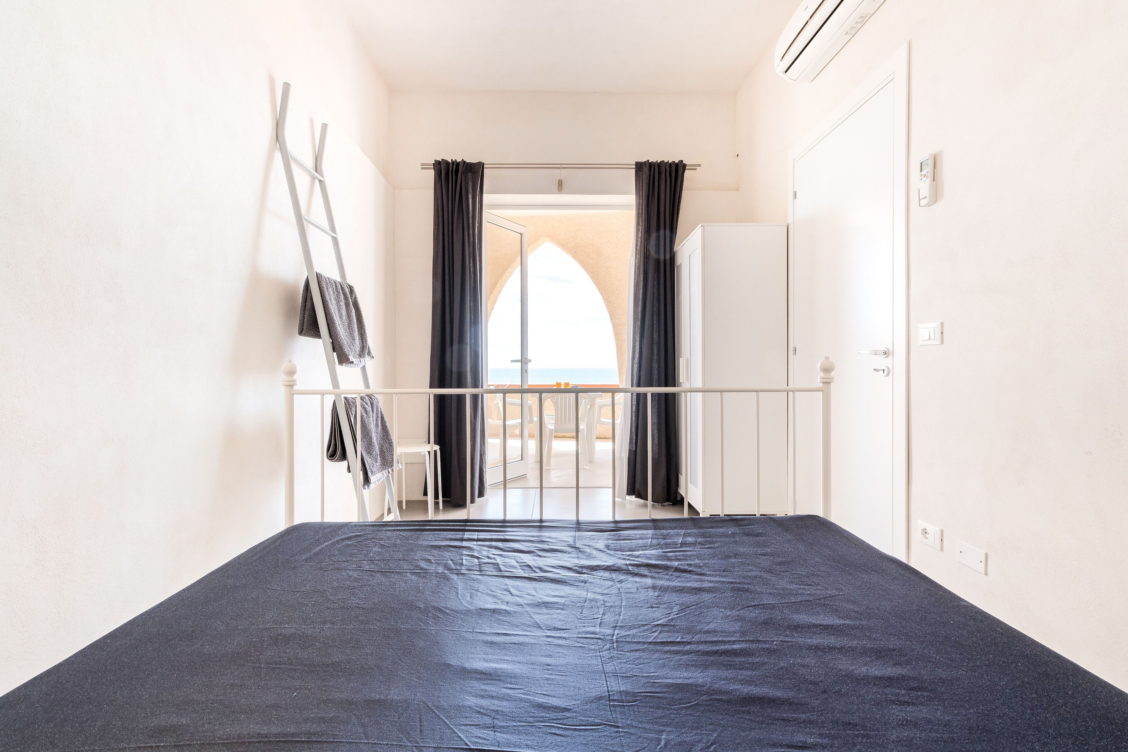 A room with a bed, a ladder, a view through an arched doorway, and a door.