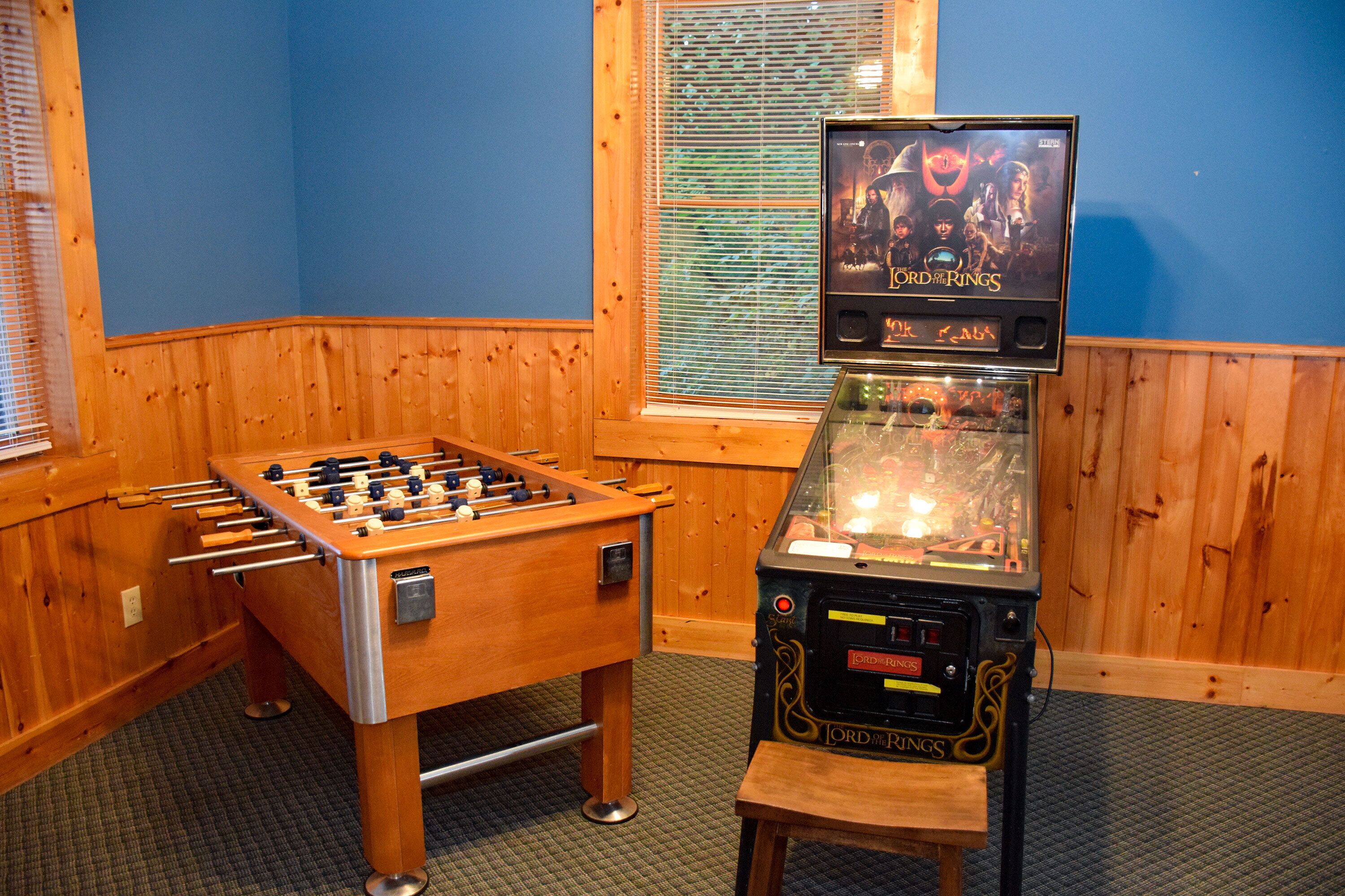 A foosball table and a video game machine in a room with wooden walls and a window with blinds.
