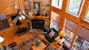 A cozy cabin interior with a stone fireplace, wooden walls, a ceiling fan, and large windows. - Hiker's Point - Cabin In The Smokies (Sevierville)