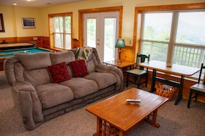 A cozy living room with a pool table, a sofa with pillows, a wooden coffee table, and a view through large windows.