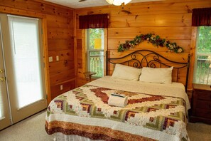 A wooden cabin bedroom with a bed, a nightstand, a window with shutters, and a door.