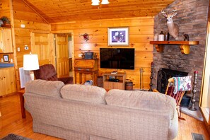 A cozy cabin interior with a large sofa, a television, a fireplace, and wooden walls.