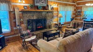 A cozy living room with a stone fireplace, wooden furniture, and a plaid-patterned chair. - Sitting Rock - Cabin In The Smokies (Sevierville)