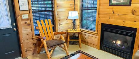 Un interior de cabaña acogedor, con una silla de madera, una mesita con una lámpara, una chimenea y un televisor montado en la pared.
